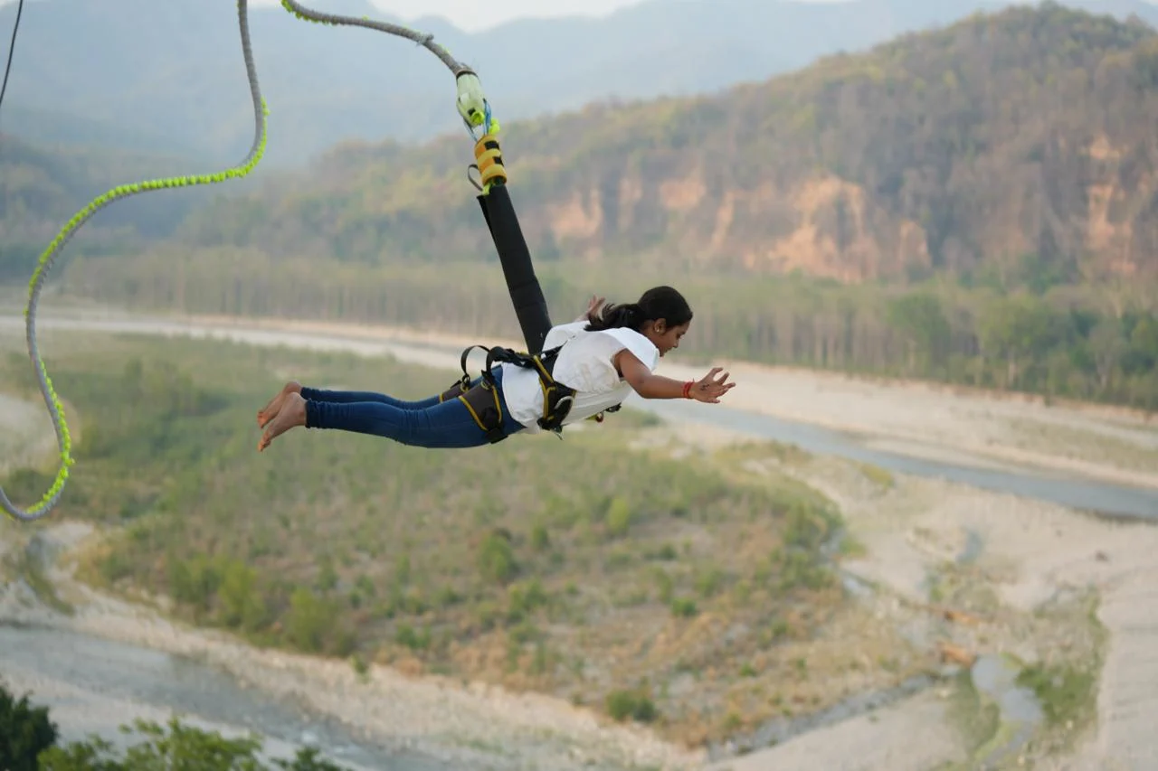 A young woman bungee jumping with arms spread wide over a river valley surrounded by green hills and mountains in Rishikesh, India.