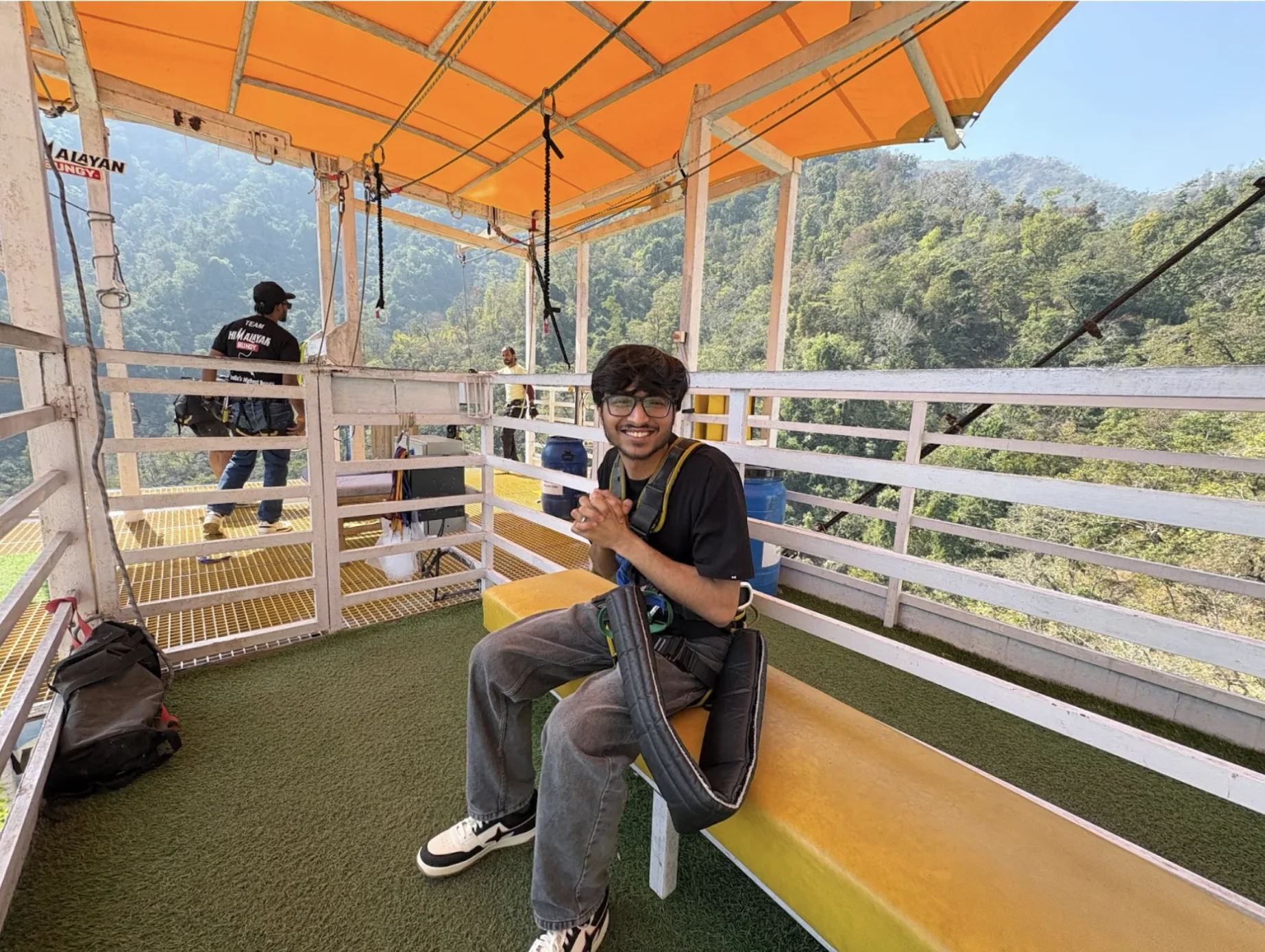 A young man smiling in a safety harness on a bungee jumping platform overlooking the green mountains of Rishikesh, India.